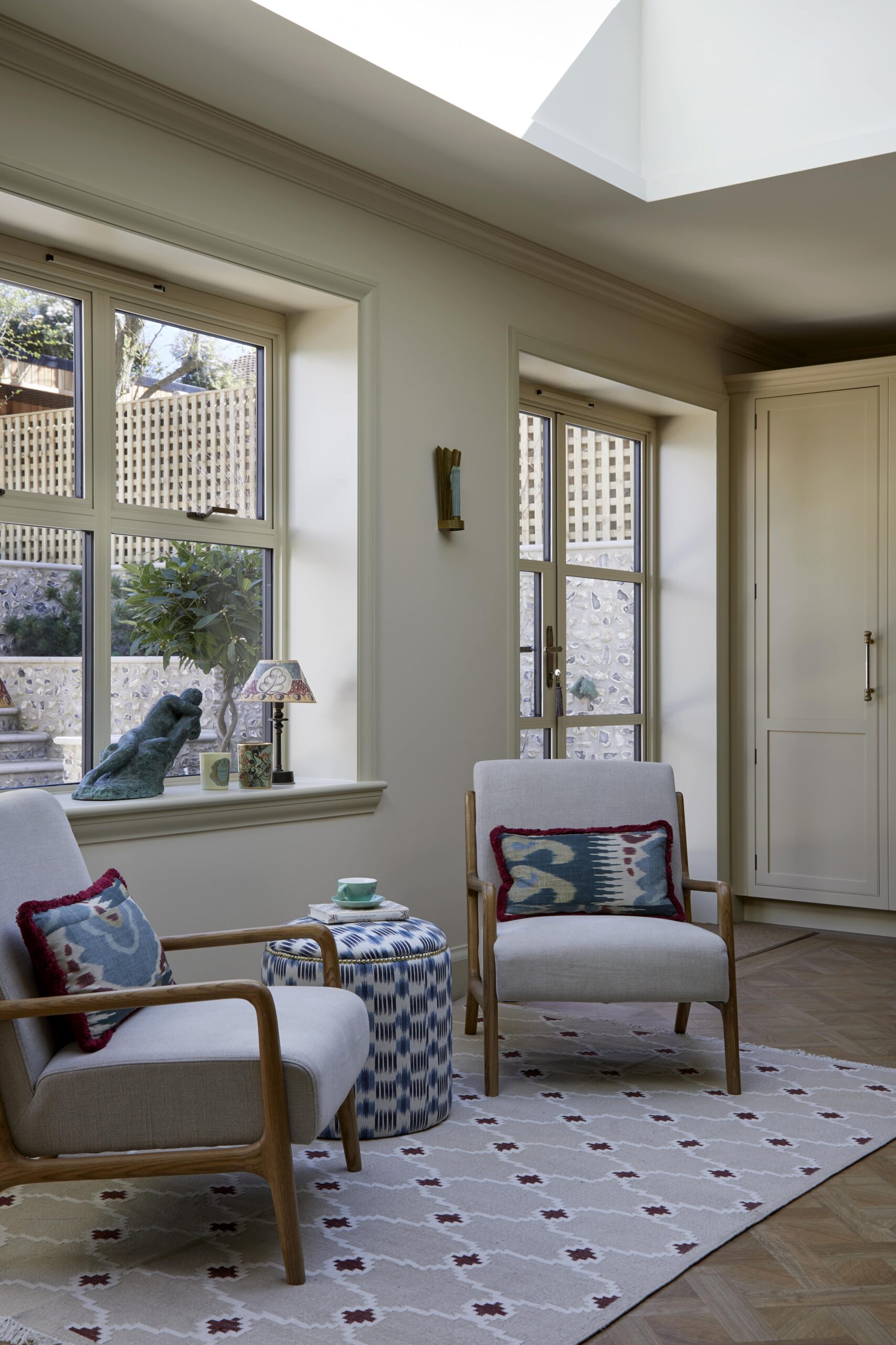Interior view of a cosy nook with two armchairs and a side table, positioned by a window with a garden view. Decor includes patterned cushions, a small rug, and tasteful ornaments on the window sill.