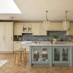 A spacious kitchen featuring yellow cabinetry with a central island in blue, topped with marble. There are elegant glass pendant lights, a parquet floor, and a cosy seating area with a patterned chair and rug.