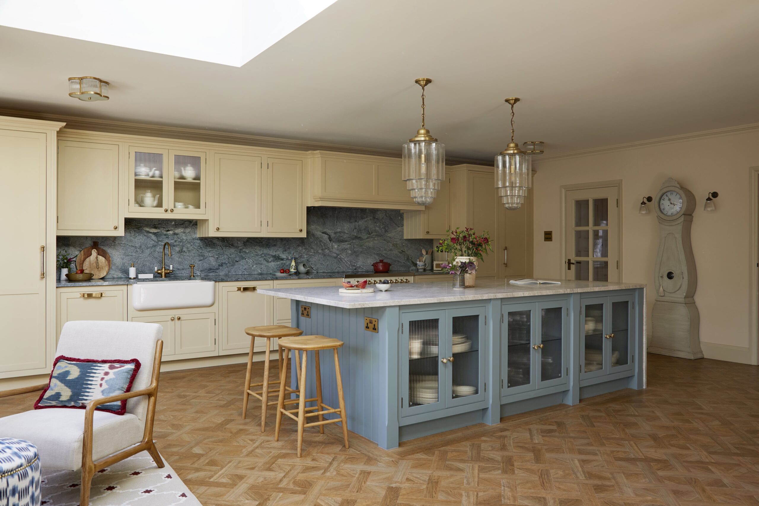 A spacious kitchen featuring yellow cabinetry with a central island in blue, topped with marble. There are elegant glass pendant lights, a parquet floor, and a cosy seating area with a patterned chair and rug.