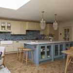 A spacious kitchen featuring yellow cabinetry with a central island in blue, topped with marble. There are elegant glass pendant lights, a parquet floor, and a cosy seating area with a patterned chair and rug.