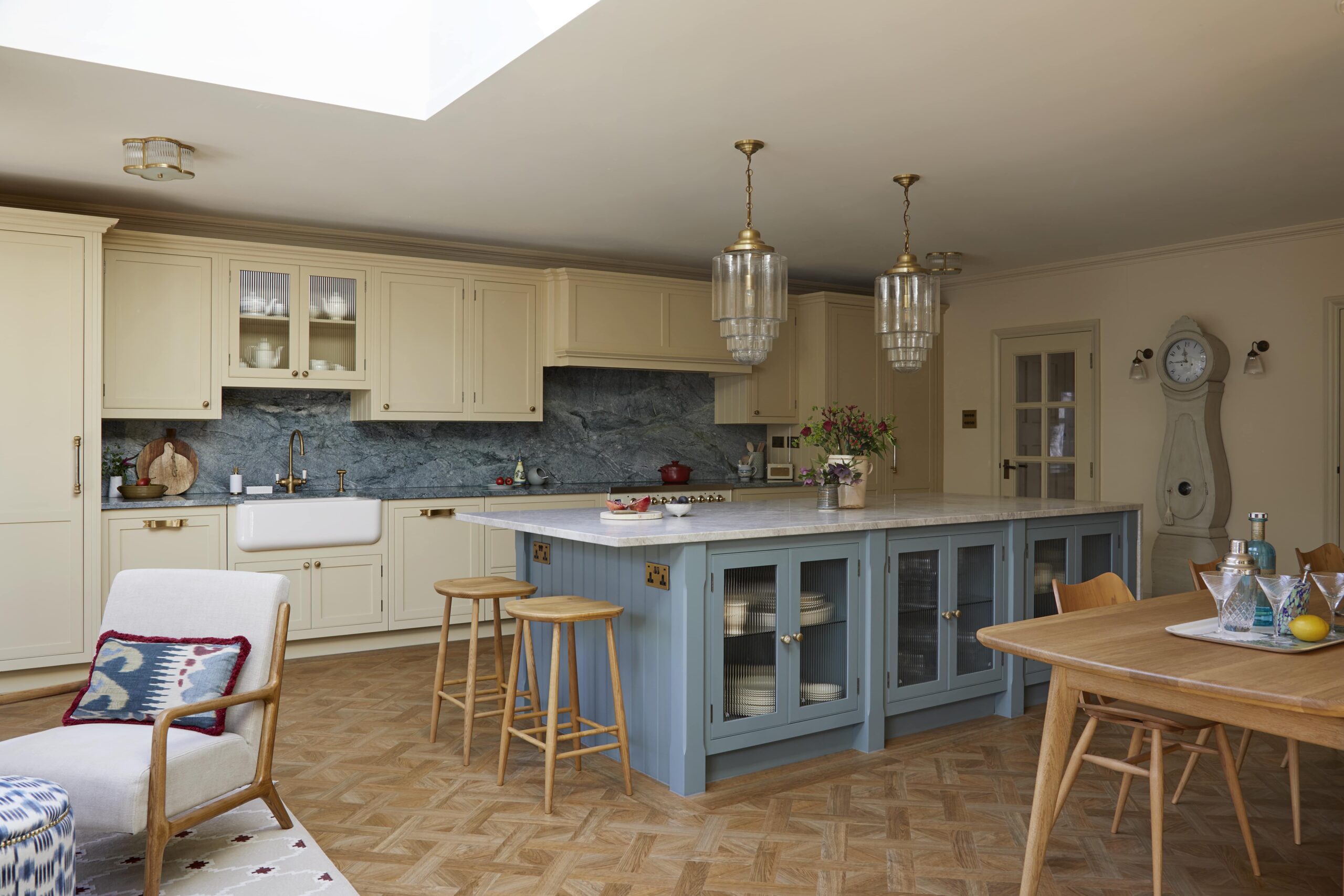 A spacious kitchen featuring yellow cabinetry with a central island in blue, topped with marble. There are elegant glass pendant lights, a parquet floor, and a cosy seating area with a patterned chair and rug.