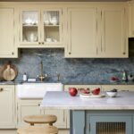 Elegant kitchen featuring yellow cabinetry with glass doors, a marble countertop, and a blue island. The backsplash is made of textured blue stone, and there's a wooden stool by the counter. Decorative items include red apples, a bowl, and a wooden cutting board.