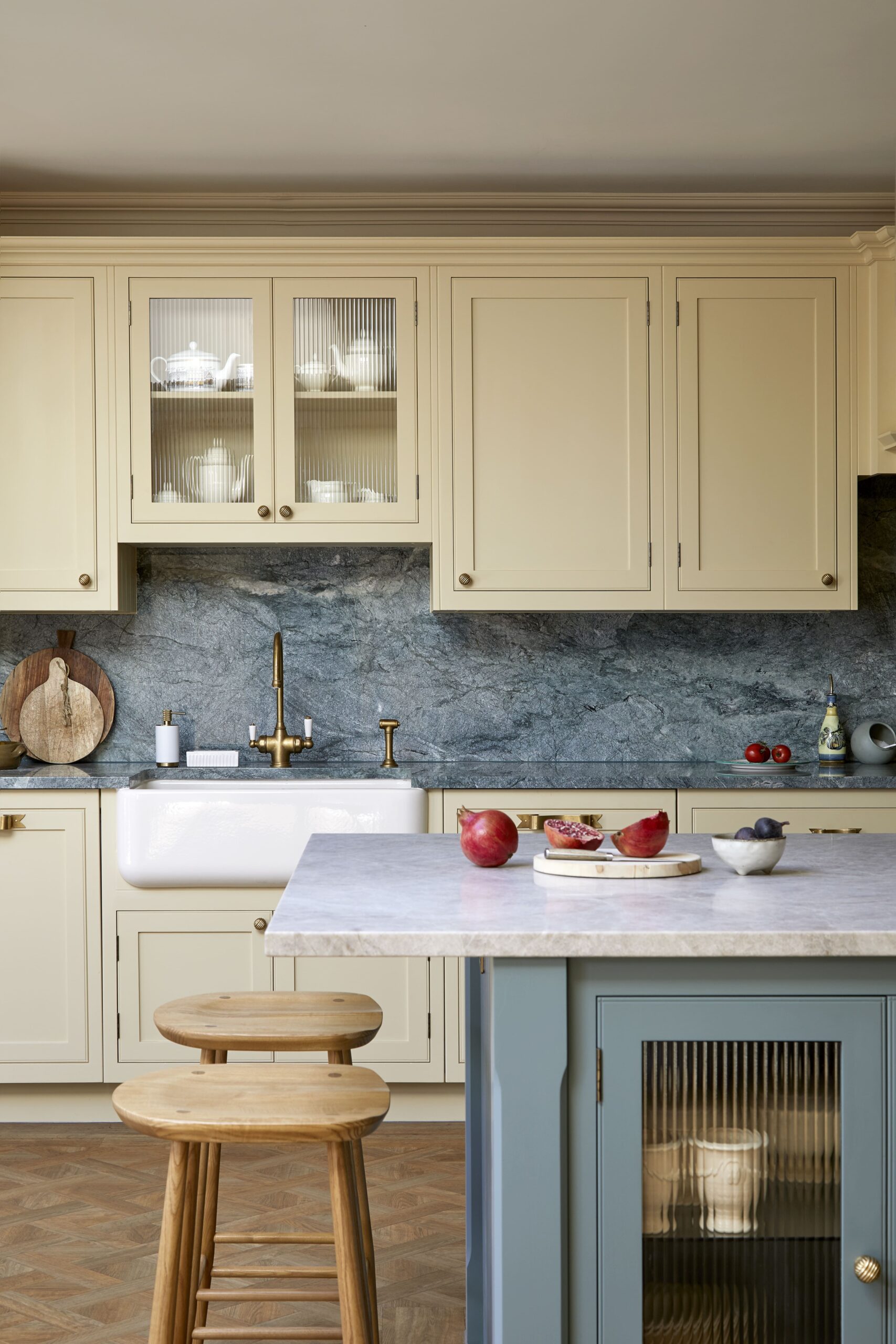 Elegant kitchen featuring yellow cabinetry with glass doors, a marble countertop, and a blue island. The backsplash is made of textured blue stone, and there's a wooden stool by the counter. Decorative items include red apples, a bowl, and a wooden cutting board.