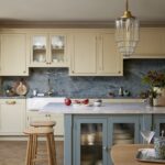 Elegant kitchen featuring yellow cabinetry with glass doors, a marble countertop, and a blue island. The backsplash is made of textured blue stone, and there's a wooden stool by the counter. Decorative items include red apples, a bowl, and a wooden cutting board.