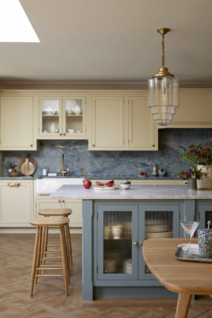 Elegant kitchen featuring yellow cabinetry with glass doors, a marble countertop, and a blue island. The backsplash is made of textured blue stone, and there's a wooden stool by the counter. Decorative items include red apples, a bowl, and a wooden cutting board.