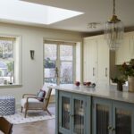Interior view of a bright kitchen featuring blue island cabinets topped with a marble countertop, and a pendant light above it. The space is complemented by a cosy seating area with a chair, a cushioned stool, and a large window allowing natural light to enhance the room.