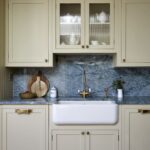 Elegant kitchen featuring soft yellow coloured cabinetry with glass upper cabinet doors, a white Belfast sink, and a blue-grey marble backsplash. A brass Perrin & Rowe tap and decorative items complete the setting.