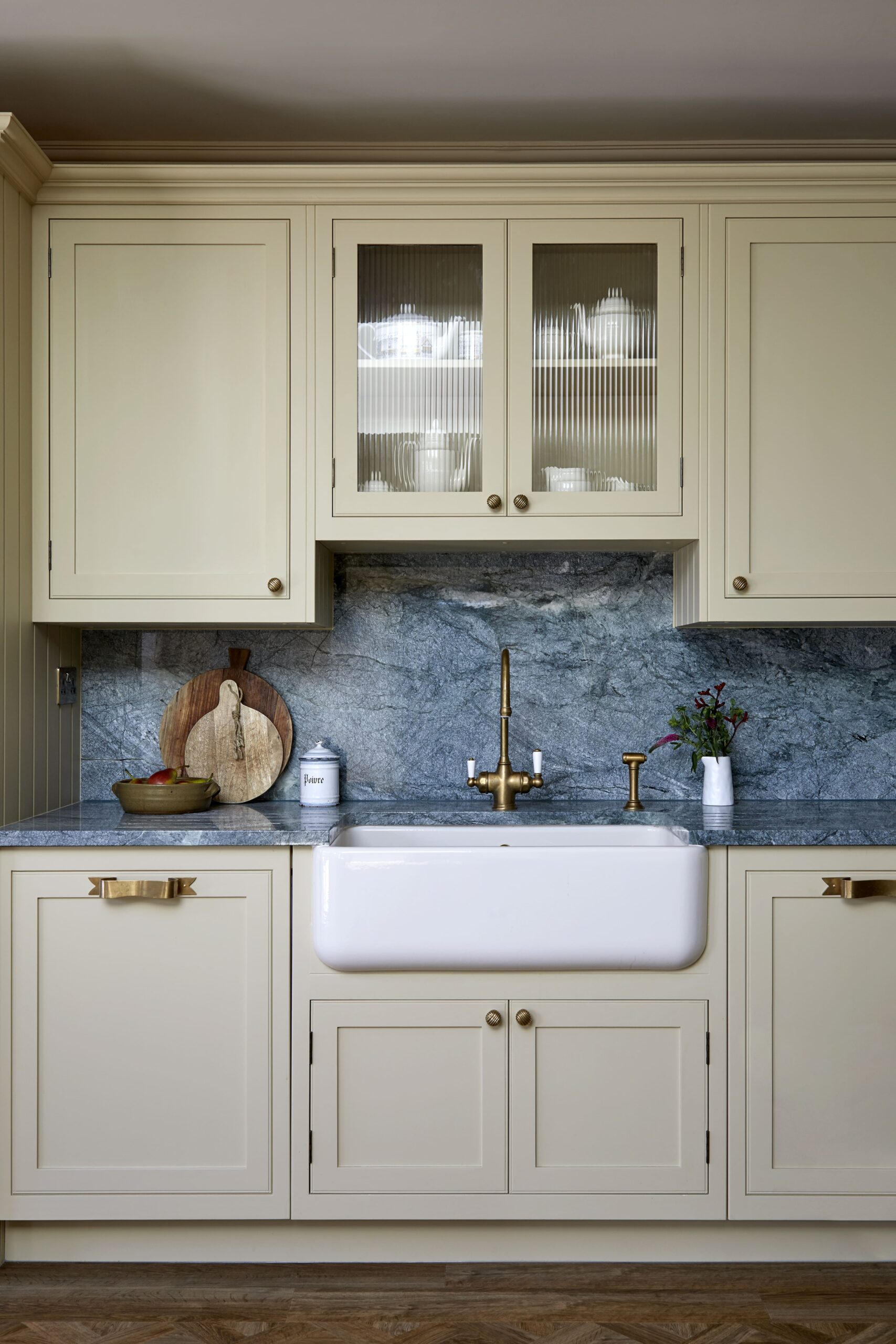 Elegant kitchen featuring soft yellow coloured cabinetry with glass upper cabinet doors, a white Belfast sink, and a blue-grey marble backsplash. A brass Perrin & Rowe tap and decorative items complete the setting.