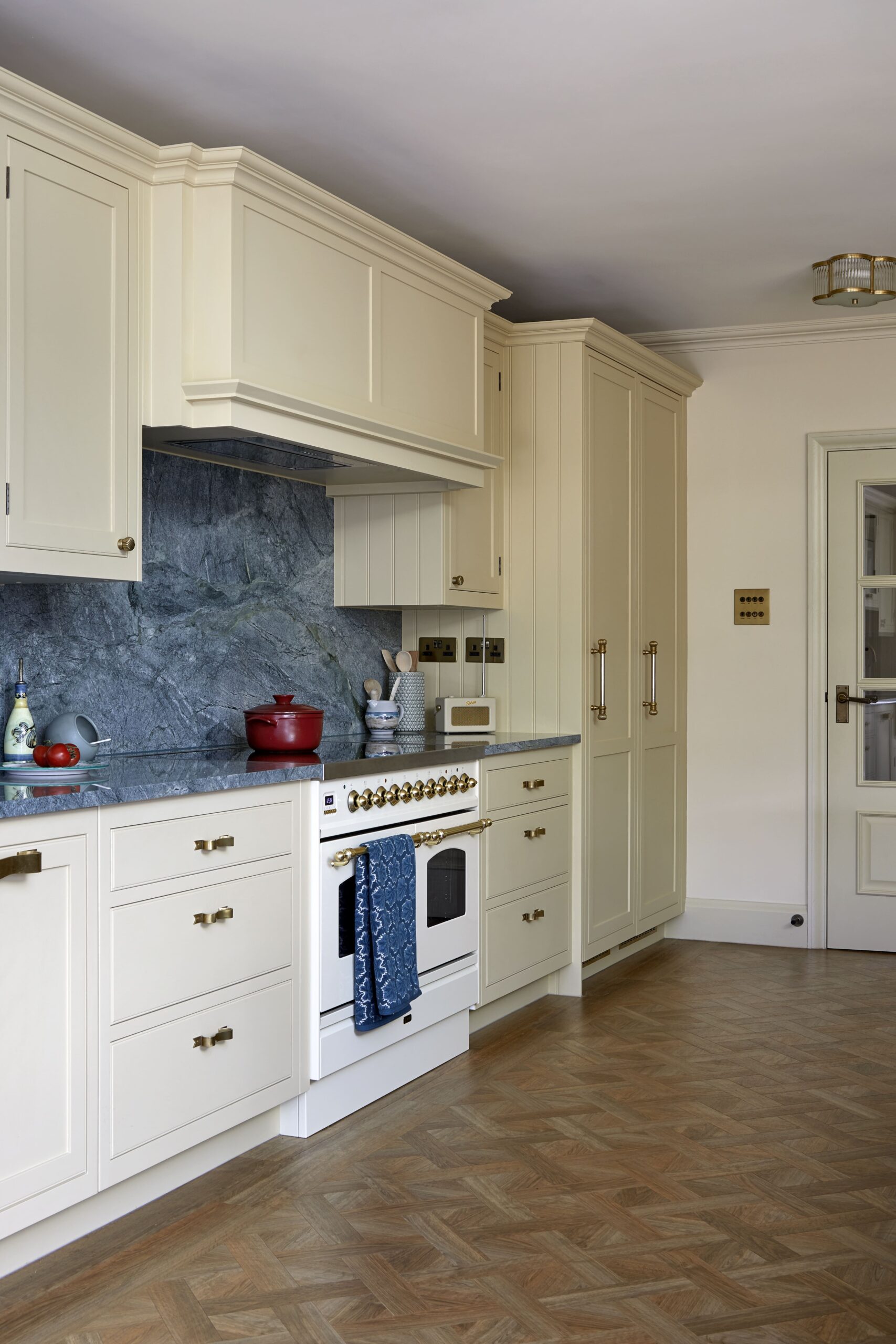 Elegant kitchen interior featuring white cabinetry, a marble backsplash, and a vintage-style stove. The room includes a herringbone patterned wood floor and a built-in refrigerator. Decorative items and cooking utensils are visible.