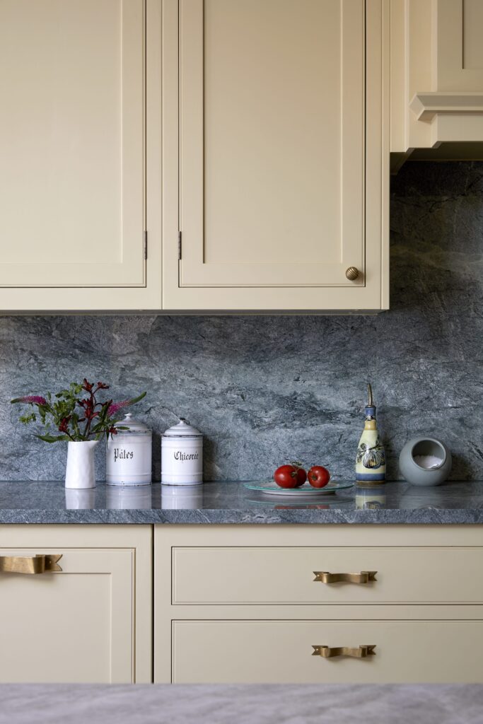 Elegant kitchen countertop with a blue marble backsplash, featuring decorative items including a floral arrangement, labelled canisters, a ceramic oil bottle, and fresh tomatoes. Light-yellow cabinetry completes the setting.