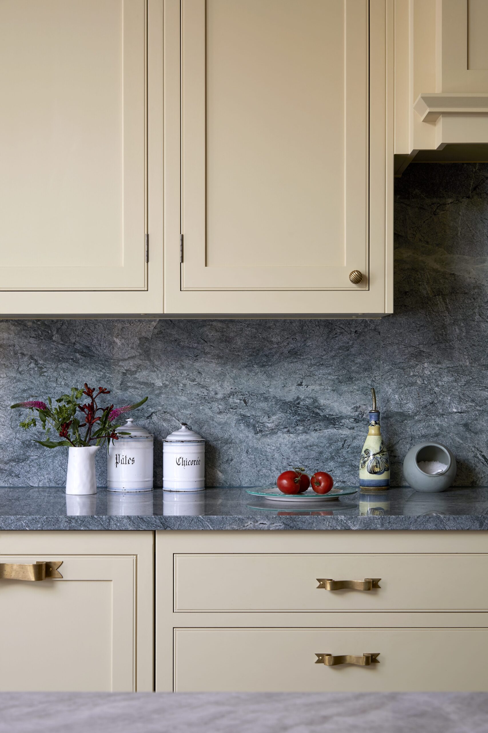 Elegant kitchen countertop with a blue marble backsplash, featuring decorative items including a floral arrangement, labelled canisters, a ceramic oil bottle, and fresh tomatoes. Light-yellow cabinetry completes the setting.