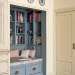 From a side angle, is a cosy built-in reading nook painted in light blue, featuring a cushioned seat with pillows, flanked by shelves filled with books and decorative items.