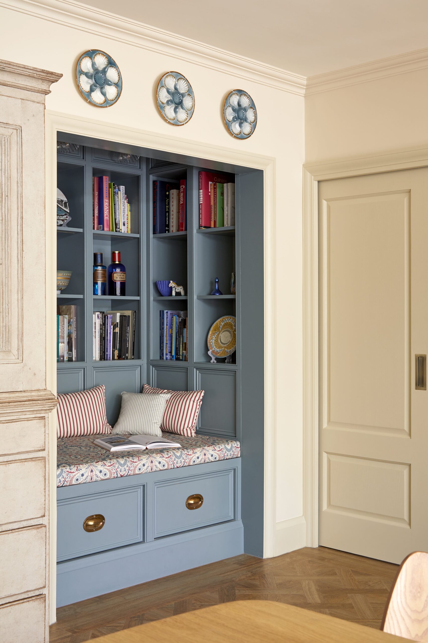 From a side angle, is a cosy built-in reading nook painted in light blue, featuring a cushioned seat with pillows, flanked by shelves filled with books and decorative items.