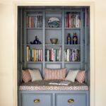 A cosy built-in reading nook painted in light blue, featuring a cushioned seat with pillows, flanked by shelves filled with books and decorative items.