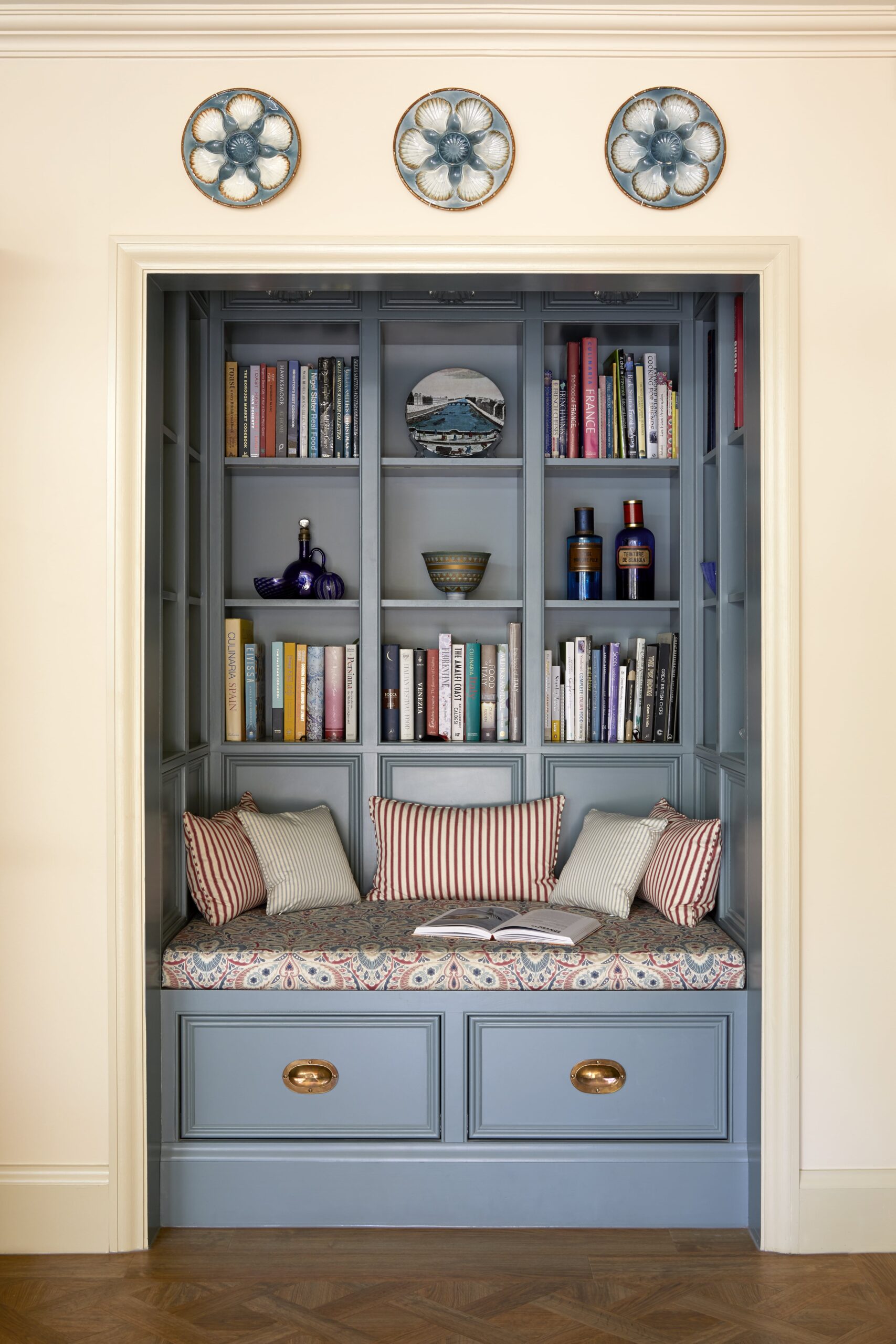 A cosy built-in reading nook painted in light blue, featuring a cushioned seat with pillows, flanked by shelves filled with books and decorative items.