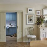 Elegant interior of a room featuring a wooden herringbone floor, pale yellow walls, and a vintage white dresser with a mirror. The room includes decorative plates on the wall and a floral arrangement on the dresser. An open door reveals a blue utility room in the background.