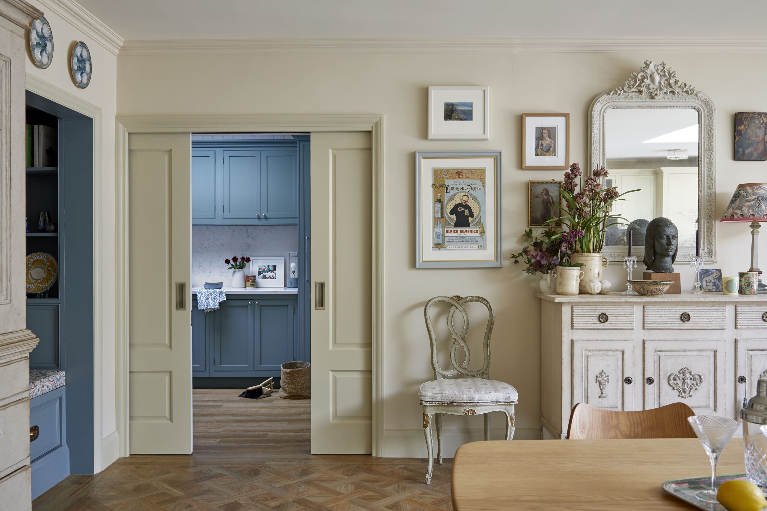 Elegant interior of a room featuring a wooden herringbone floor, pale yellow walls, and a vintage white dresser with a mirror. The room includes decorative plates on the wall and a floral arrangement on the dresser. An open door reveals a blue utility room in the background.