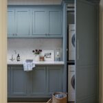 A tidy utility room featuring blue cabinets, a stacked washing machine and dryer, a small sink, and a wicker basket with a pair of shoes beside it.
