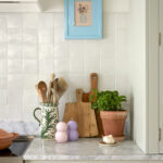 A bespoke kitchen, personalisation displays a carefully curated countertop, where a wooden bowl, matching chopping and cutting boards, and a floral mug create a harmonious blend of nature and craftsmanship. The white tiles and blue picture frame reflect a unique touch, turning this space into a warm and inviting oasis for every cook.