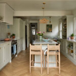 A indoor outdoor kitchen, kitchen island displays a harmonious blend of modernity and rusticity. The island, with its white marble and wooden countertop, centerpiece, holds a blue vase and a green plant, creating a serene atmosphere. Equipped with a silver stove and microwave, the island is flanked by sleek white cabinets and a refrigerator, while a wooden bar and stool add warmth. This unique fusion of elegance and charm defines the space.
