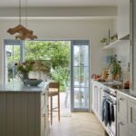 A indoor outdoor kitchen, family kitchen displays a harmonious blend of modern and rustic charm. The spacious island, with its white countertop and wooden bar, invites relaxation and connection. The garden view through the large window enhances the cozy, family-friendly atmosphere, making this a truly unique and inviting space.