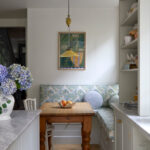 A kitchen dining, family kitchen displays a warm, inviting space where a wooden table centerpiece, adorned with fruit, invites family gatherings. The blue and white wooden bench, modern cabinets, and artistic painting create a cohesive, well-designed haven for shared meals.