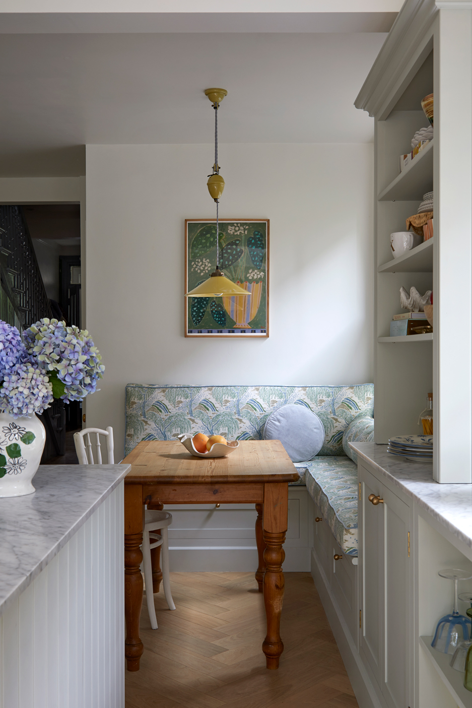 A kitchen dining, family kitchen displays a warm, inviting space where a wooden table centerpiece, adorned with fruit, invites family gatherings. The blue and white wooden bench, modern cabinets, and artistic painting create a cohesive, well-designed haven for shared meals.