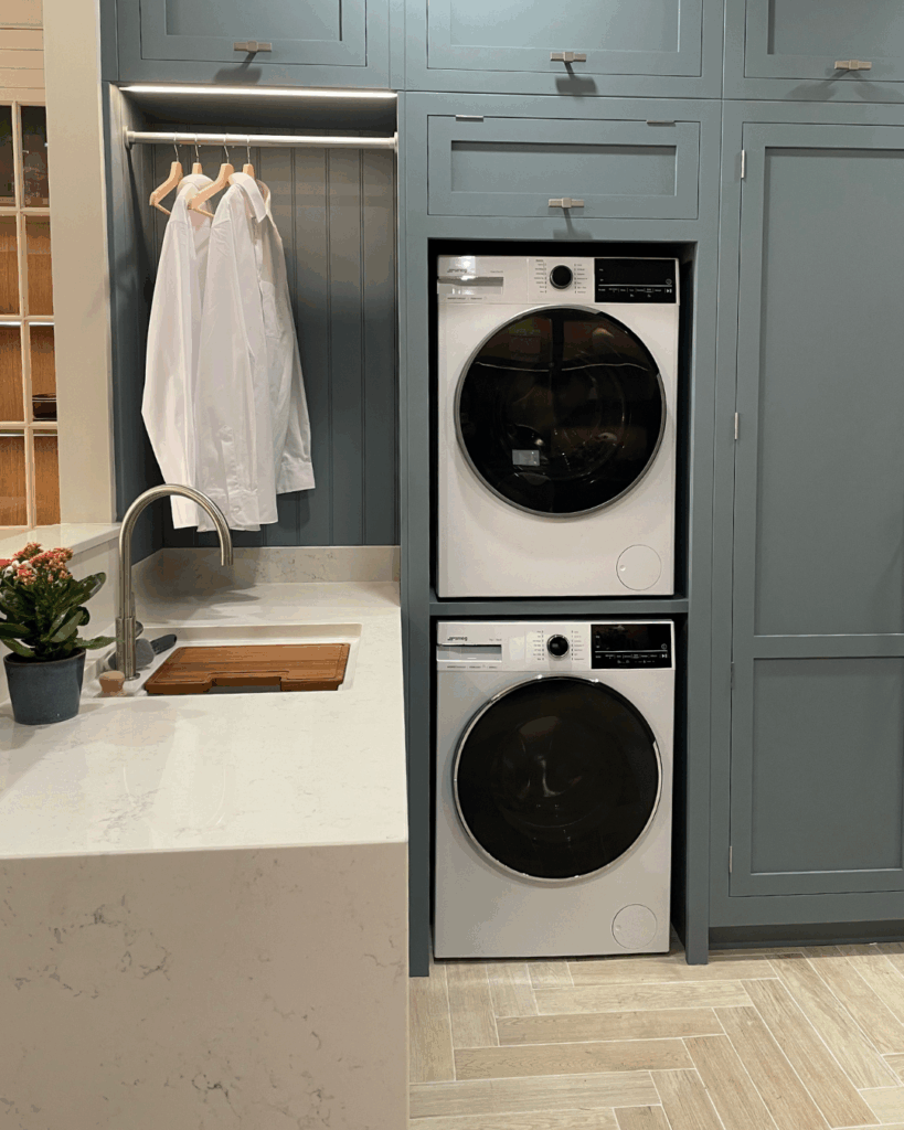 A laundry room with a stacked white washer and dryer set built into blue cabinetry, next to a sink with a white countertop. There are shirts hanging in an open cabinet above the washer.