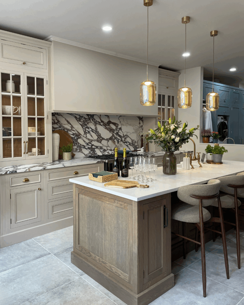 A beautiful kitchen featuring an oak central island with a marble countertop, surrounded by high stools. The space includes neutral cabinetry, built-in glass cabinets, and pendant lights. Decorations include a vase of flowers and several wine glasses.