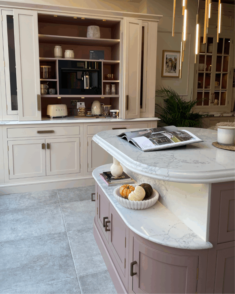 A pastel pink kitchen featuring a marble-top island with pumpkins, an open book, and a cabinet with built-in coffee machine and shelves.