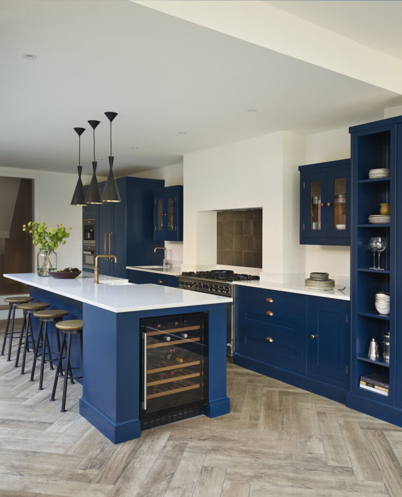 A modern kitchen featuring navy blue cabinets, a central island with bar stools, and high-end appliances including an integrated wine fridge. The flooring is light wood, and the room is well-lit with natural light.