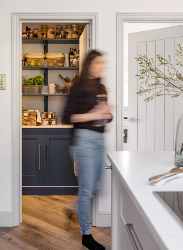 Woman walking out of walk-in pantry, designed with open shelves and classic cabinet storage, finished in navy blue.