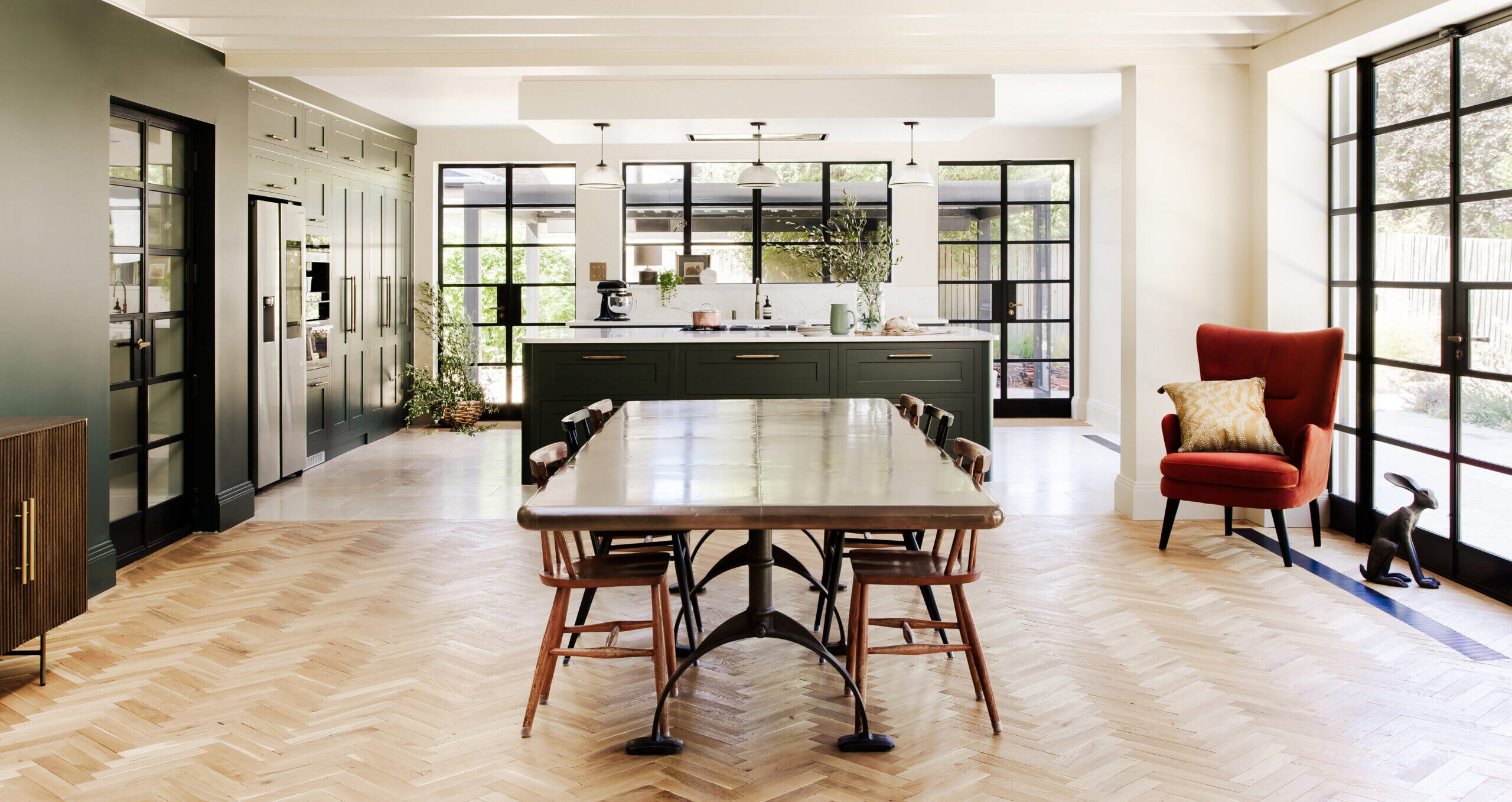 Interior view of a spacious shaker kitchen with modern design featuring a central dining table, white walls, herringbone wooden flooring, and abundant natural light from surrounding Crittall windows.