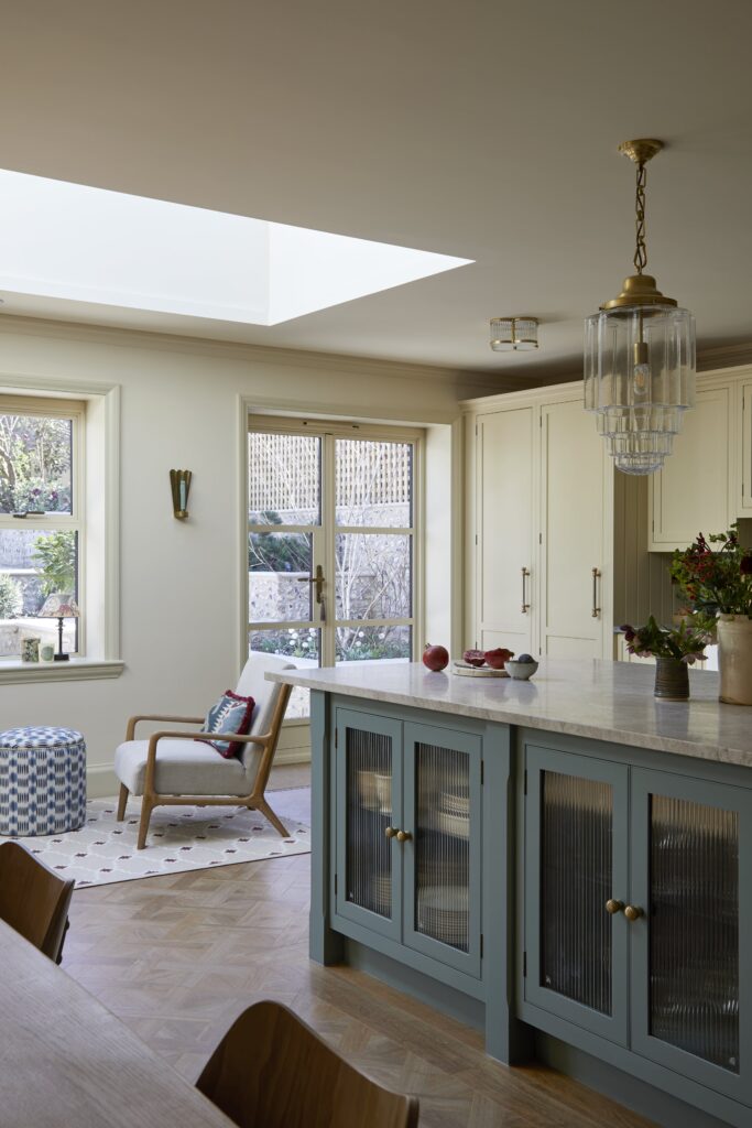 An elegant kitchen featuring a blue island with reeded glass cabinets, a light marble countertop, and a hanging glass chandelier. There's a cosy seating area by the window with a white armchair and patterned ottoman. The room has herringbone wooden floors and is well-lit with natural light from large windows.