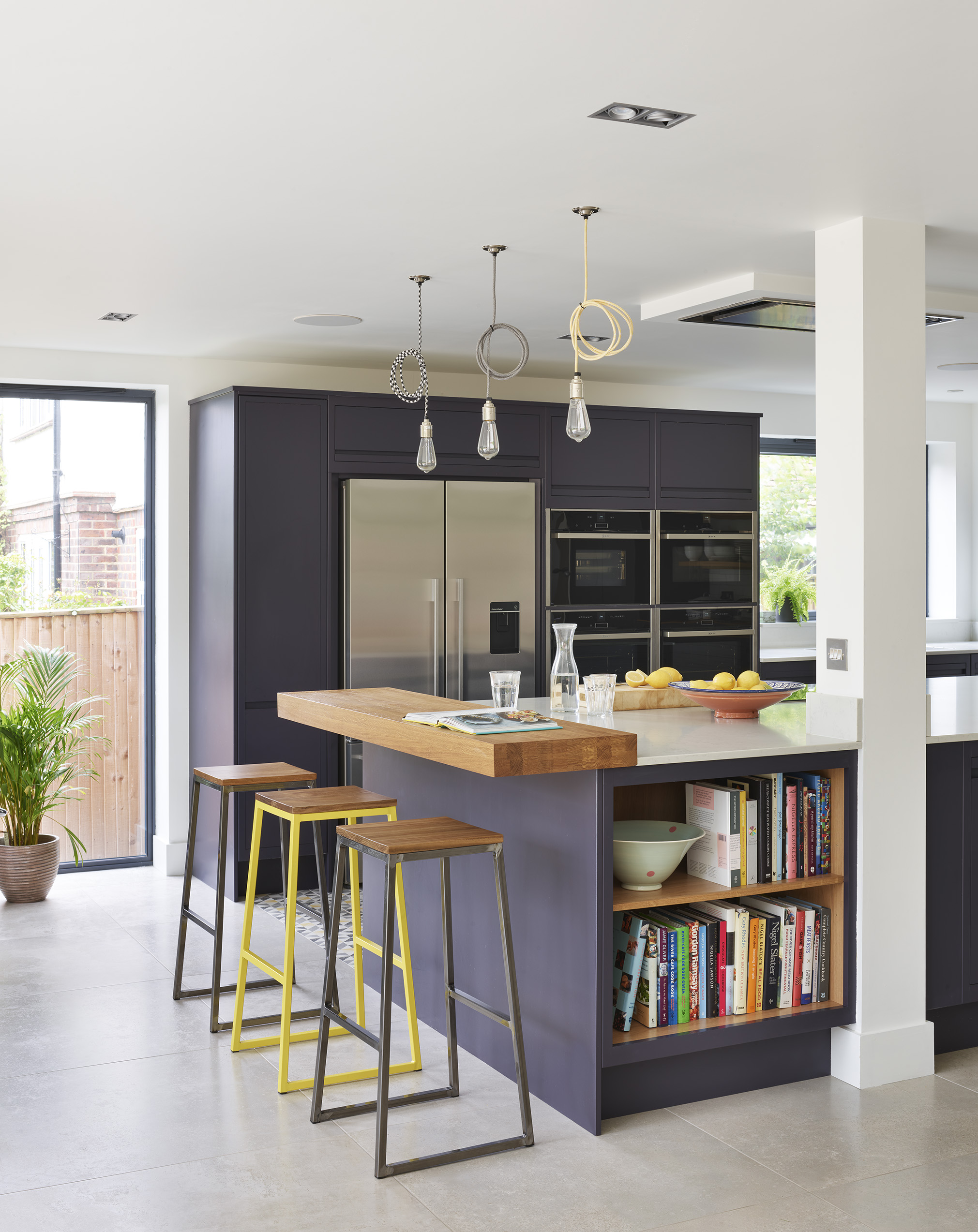 Modern navy island kitchen with quartz worktop, black and yellow bar stools and industrial style lighting.