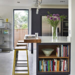 Full height windows beside a navy kitchen island with open book storage and seating in a light filled open plan space.