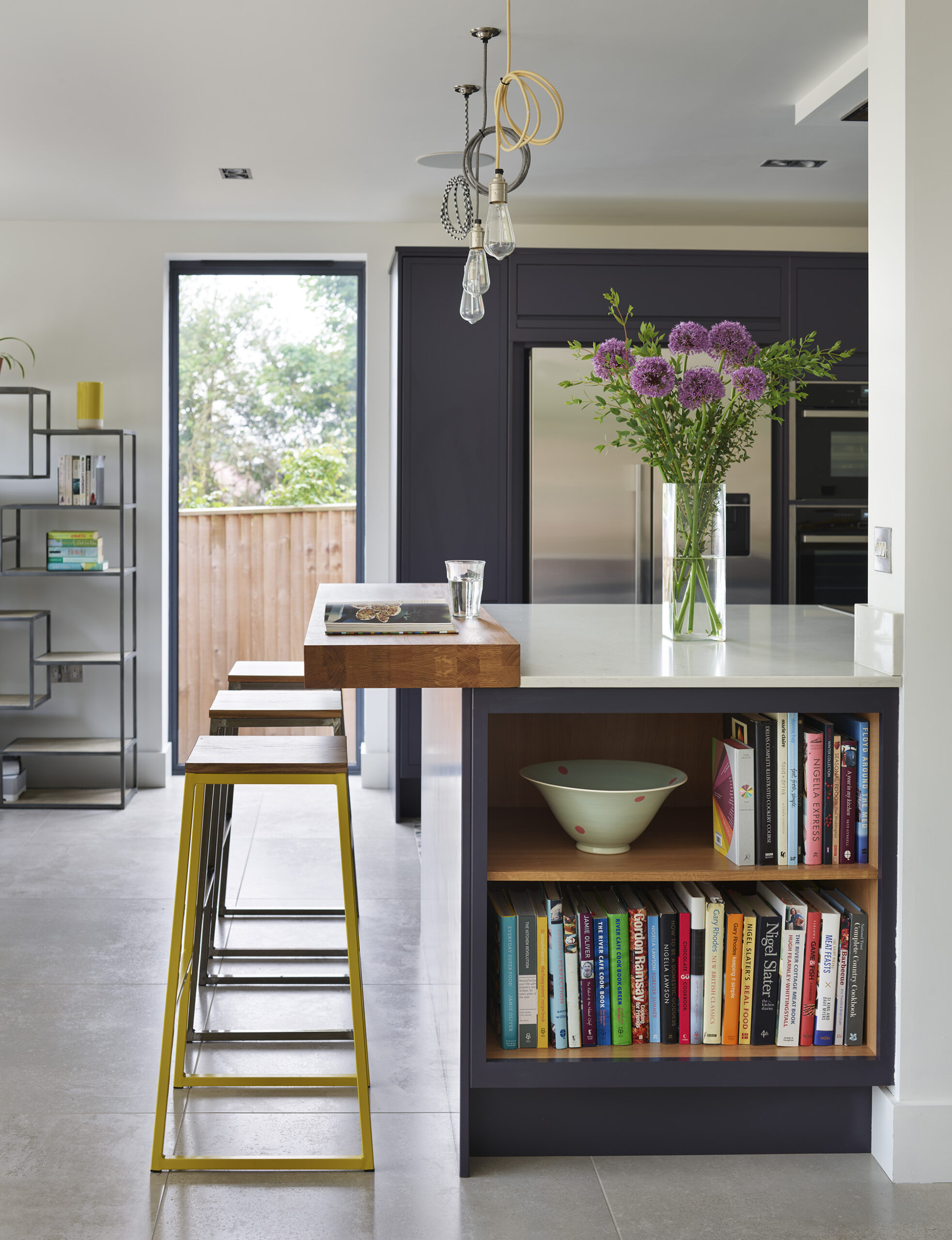 Full height windows beside a navy kitchen island with open book storage and seating in a light filled open plan space.
