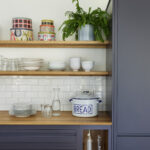 Open oak shelving in a modern navy kitchen, styled with glassware and white tiles for contrast.