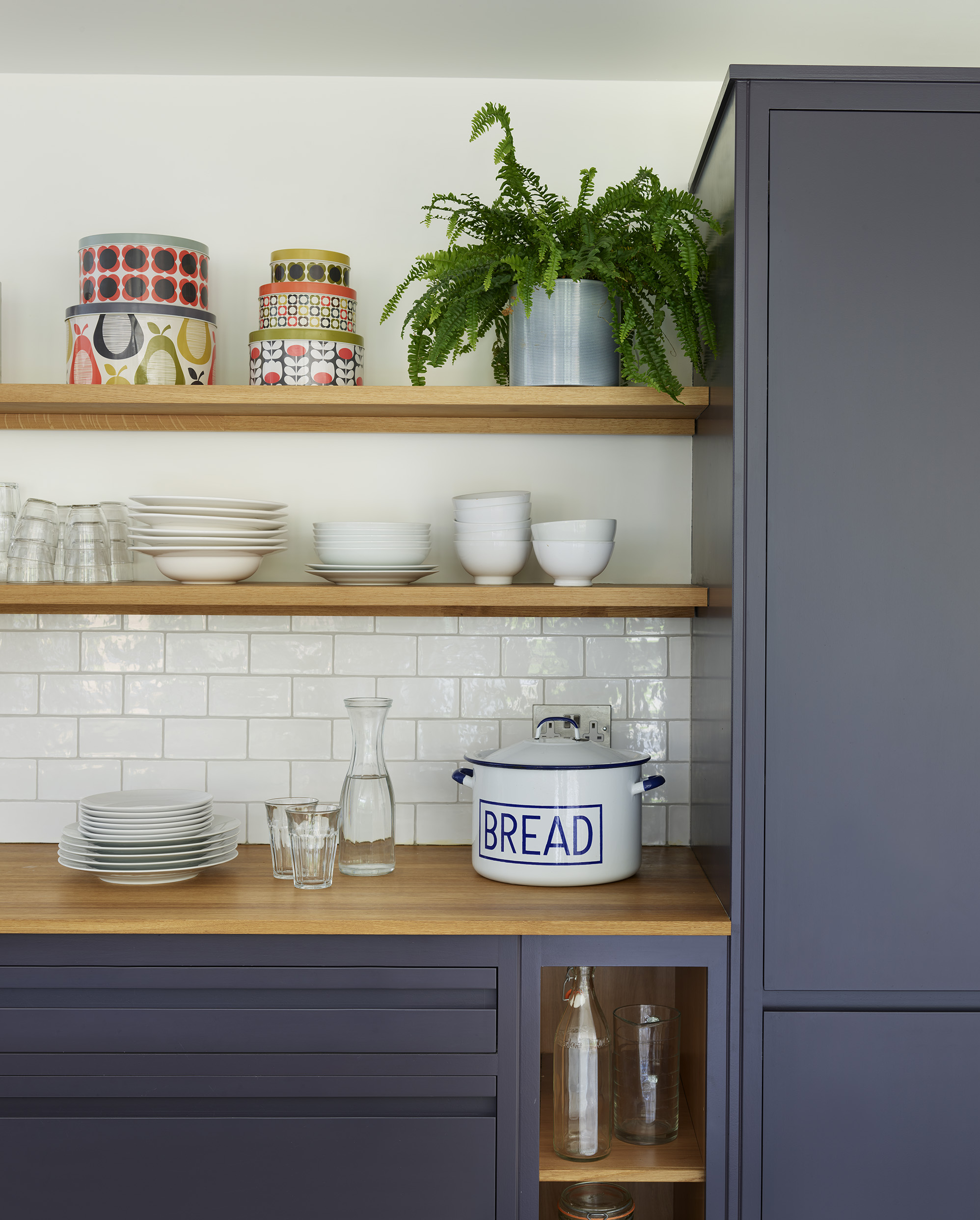 Open oak shelving in a modern navy kitchen, styled with glassware and white tiles for contrast.