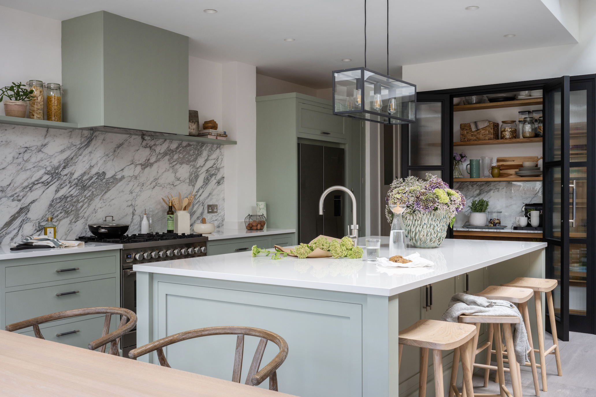 Light green modern kitchen with a large kitchen island with natural wood accents and black accents and pantry storage zone